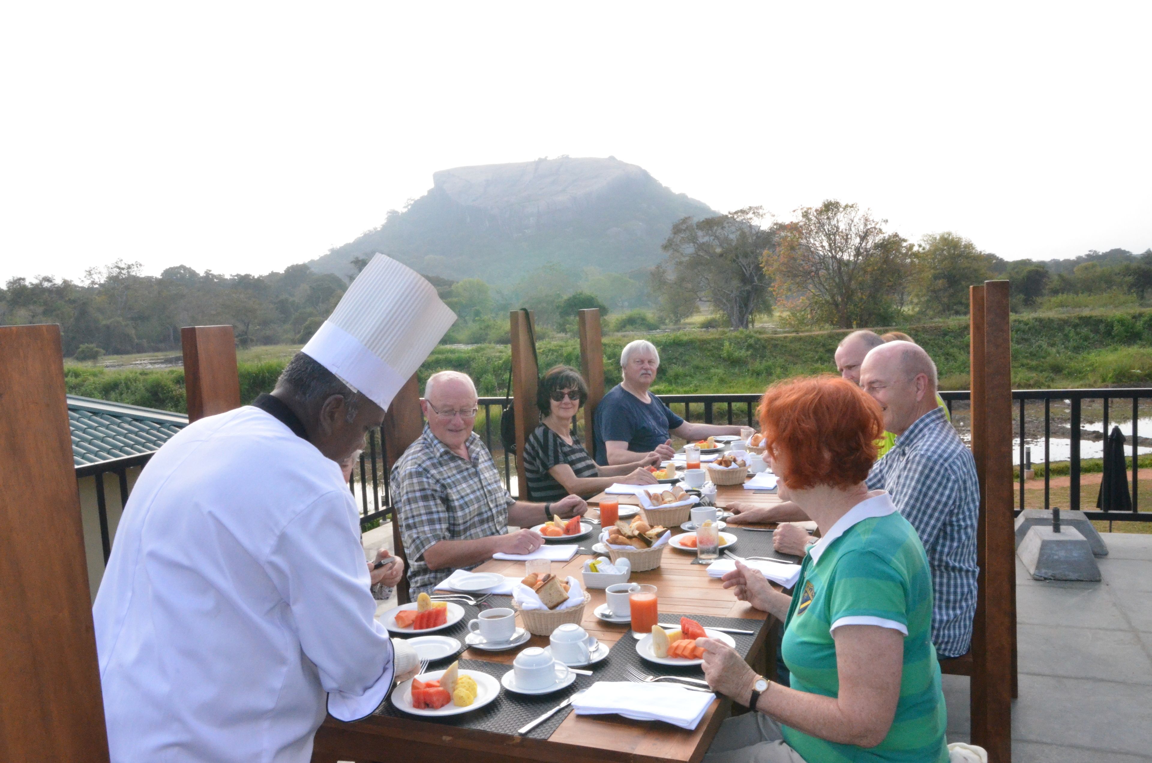 A group of people seated at a long wooden table outdoors with a scenic view of a hill in the background.
