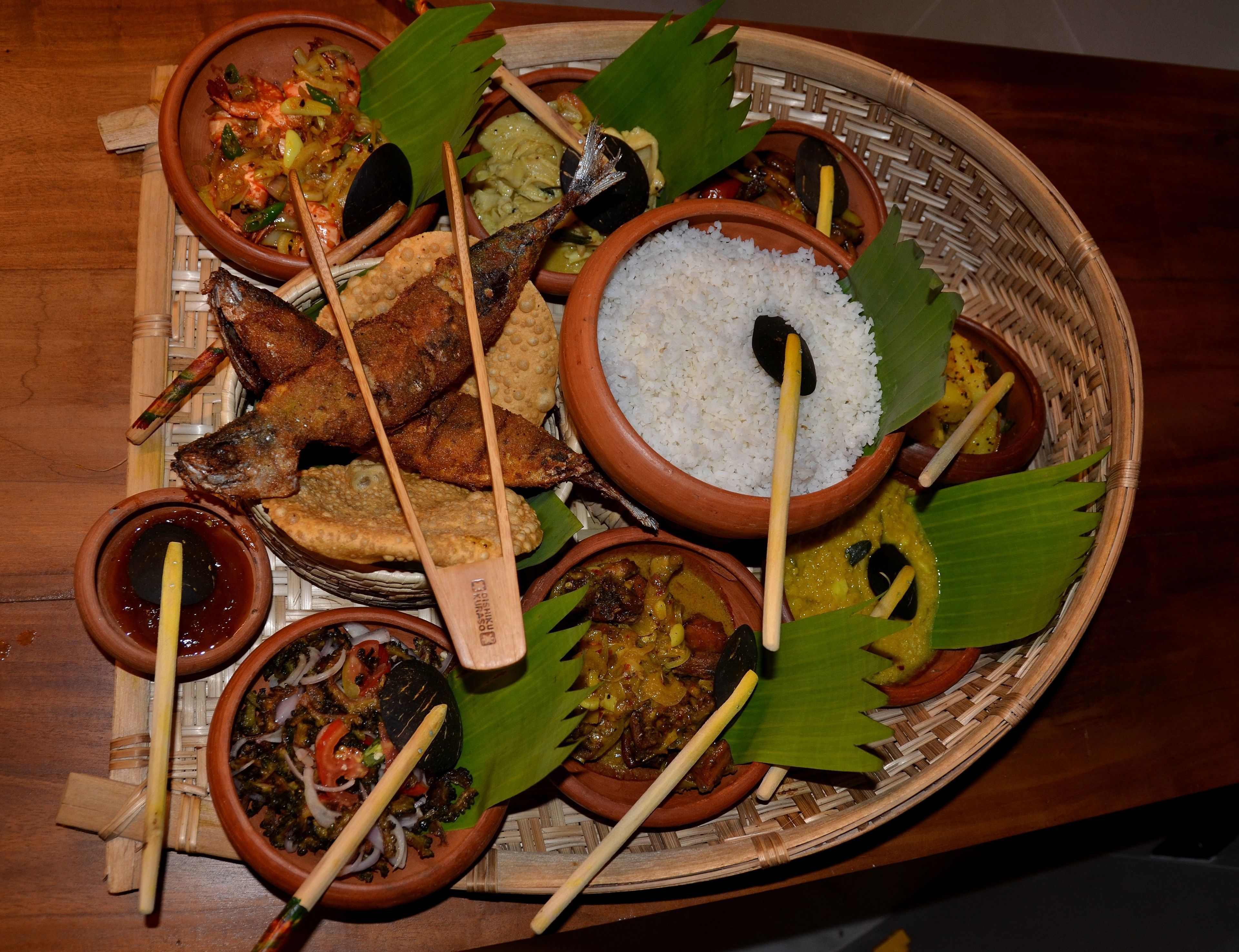 Una variedad de platos servidos en una cesta tejida, incluyendo arroz, pescado y otras preparaciones diversas, junto con utensilios de madera.