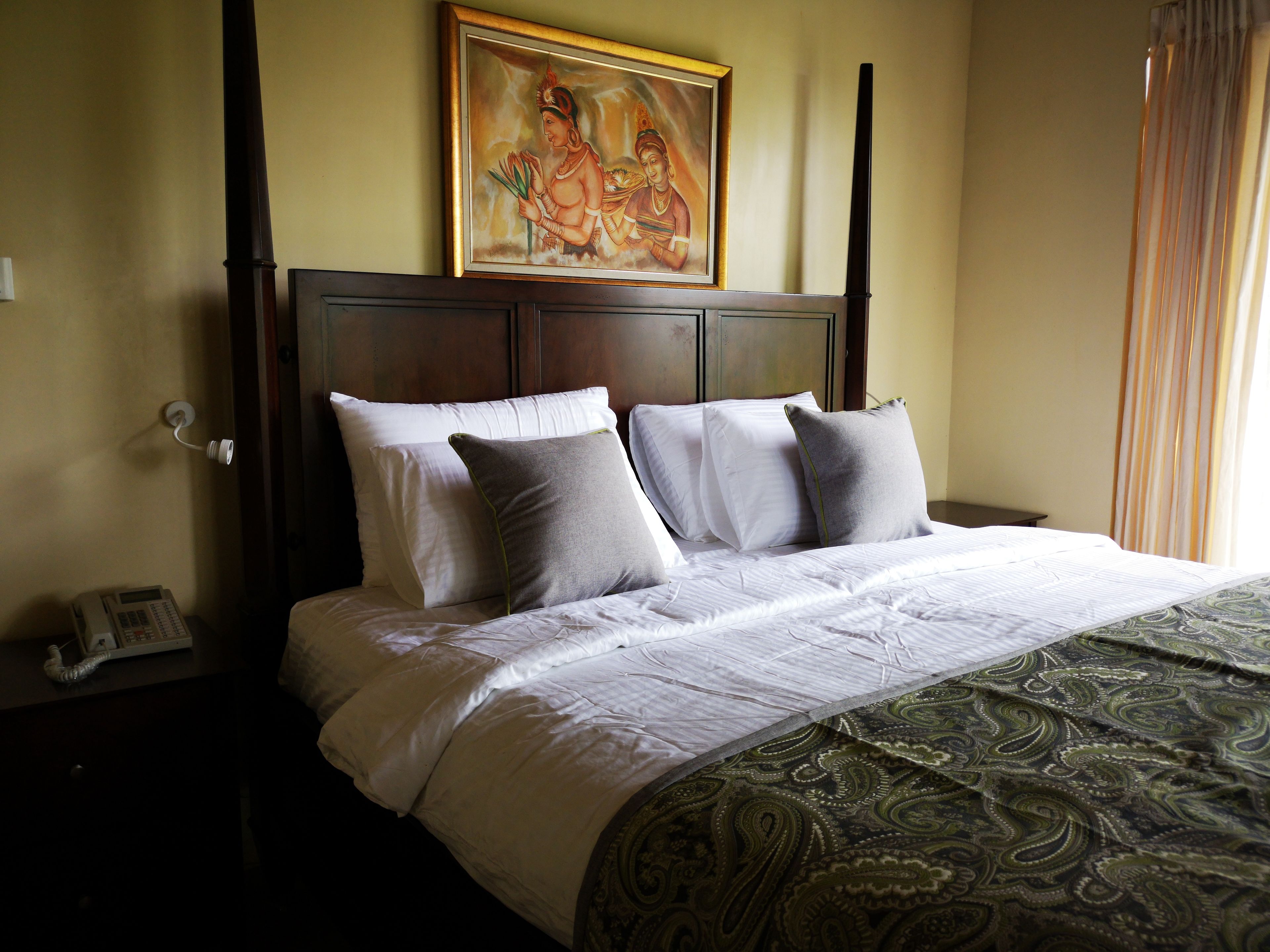 A neatly made bed with white linens and grey pillows, a patterned throw blanket, and a painting above the headboard.