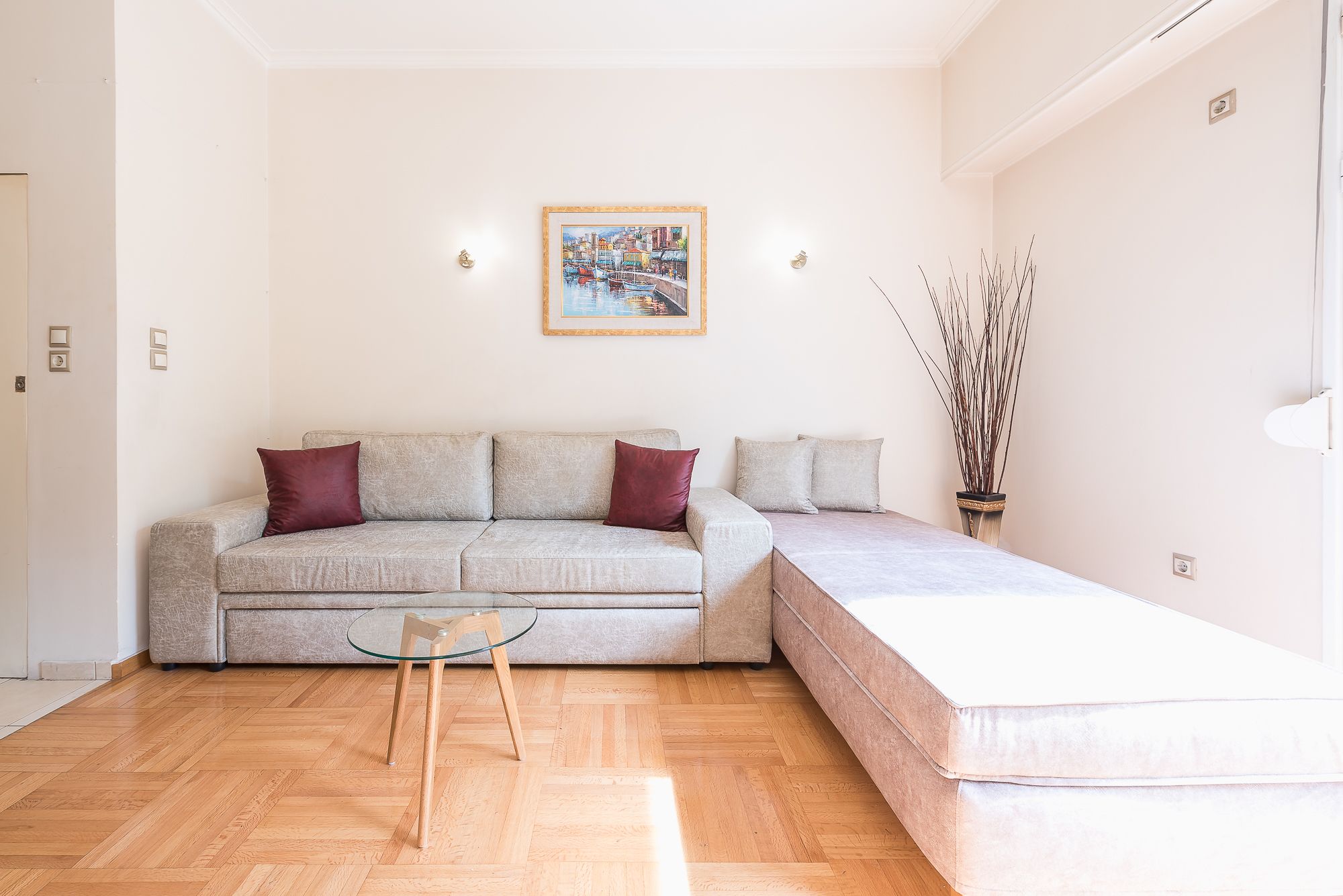 Uma sala de estar com sofá, mesa de centro de vidro, mesa lateral, um quadro na parede e um vaso com galhos.