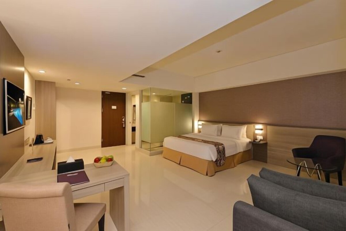A modern hotel room with a bed, bedside table, chair, desk, and a small table with fruits.