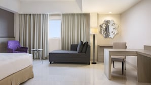 A hotel room with a bed, a sofa, a desk, a chair, a lamp, and a decorative wall piece.