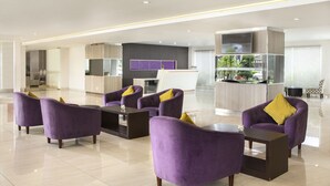 A modern hotel lobby with purple armchairs, a central coffee table, and a kitchen area in the background.