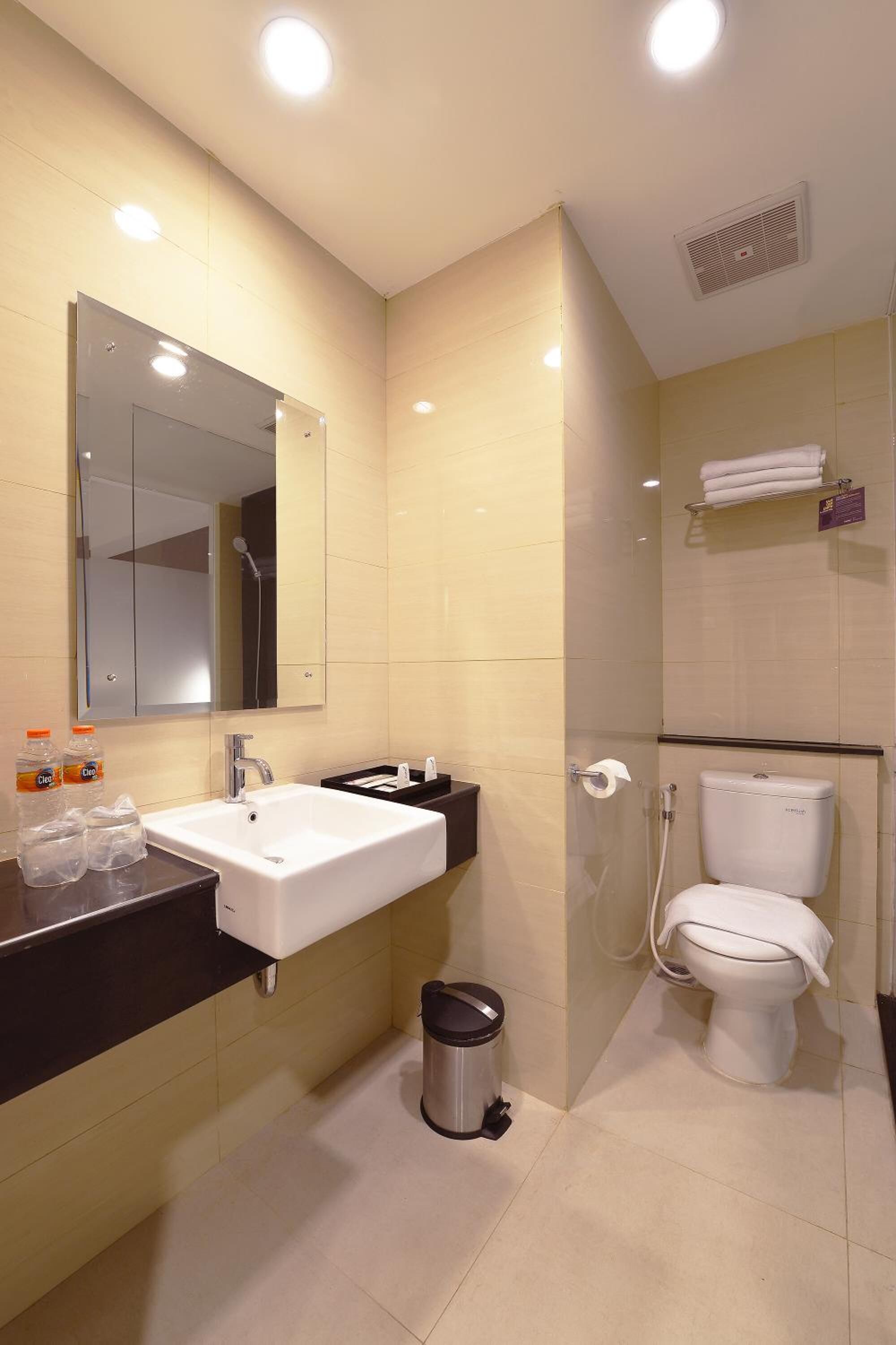 Un baño moderno con lavamanos blanco, espejo y váter. Hay una zona de ducha con puerta de vidrio, y se ve un cubo de basura.