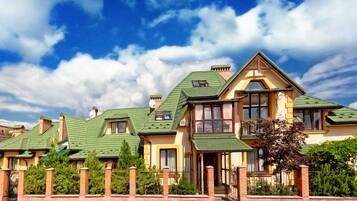 A large, multi-story house with a green roof, multiple chimneys, and a well-maintained garden.