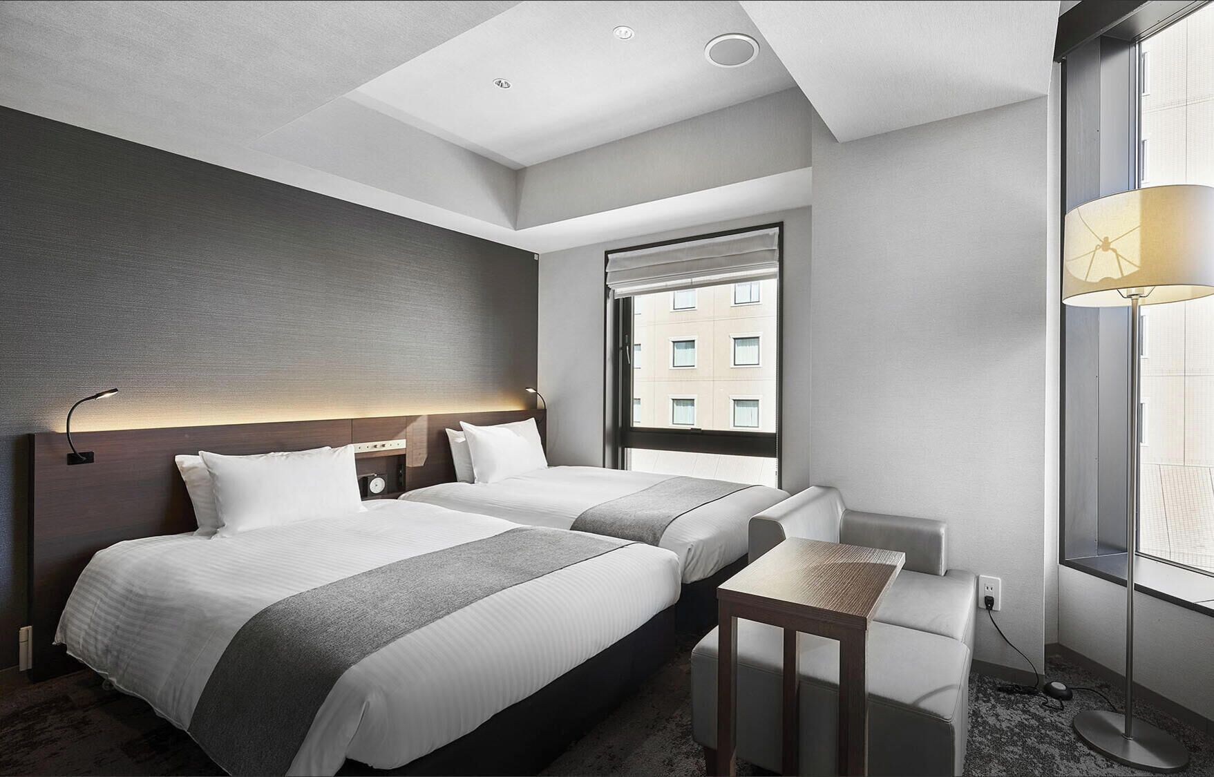 Une chambre d’hôtel avec deux lits, une petite table, une lampe et une fenêtre donnant sur des immeubles.