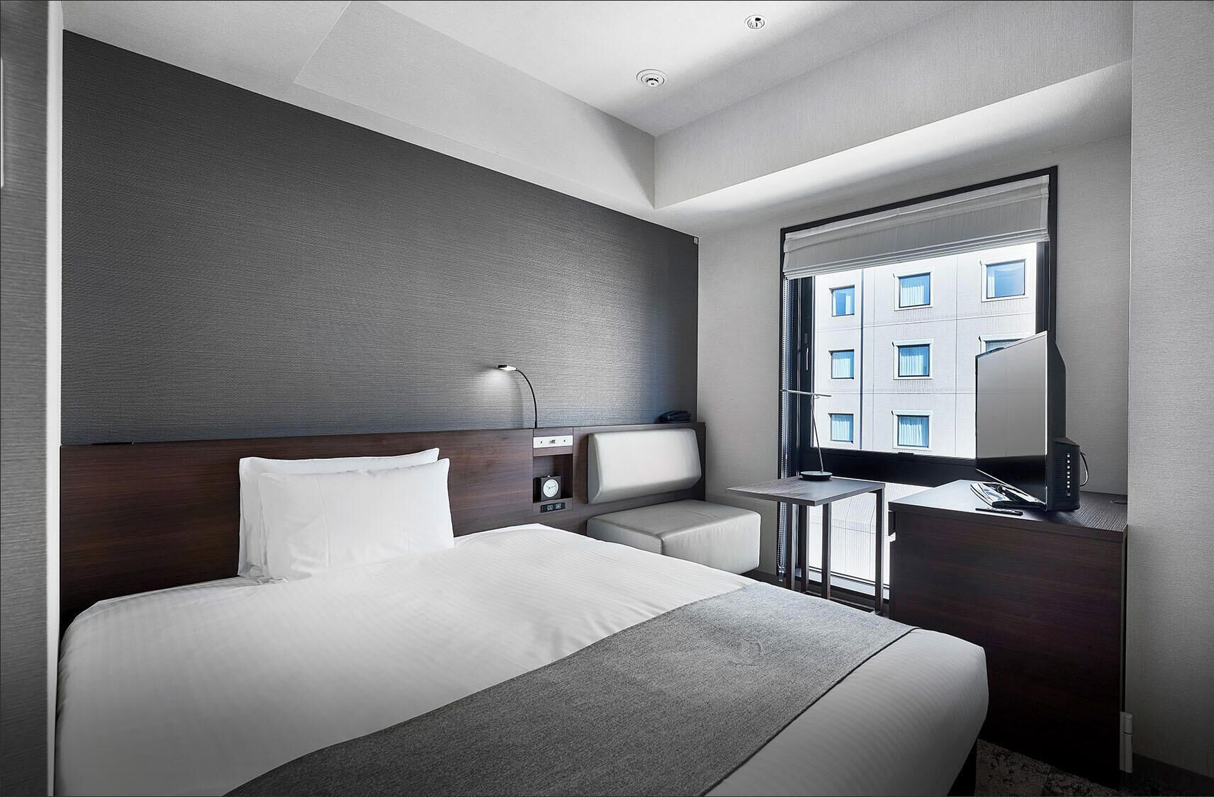 Une chambre d’hôtel moderne dotée d’un grand lit, d’un bureau, d’une chaise, d’une table de chevet, d’une lampe et d’une fenêtre donnant sur des immeubles.