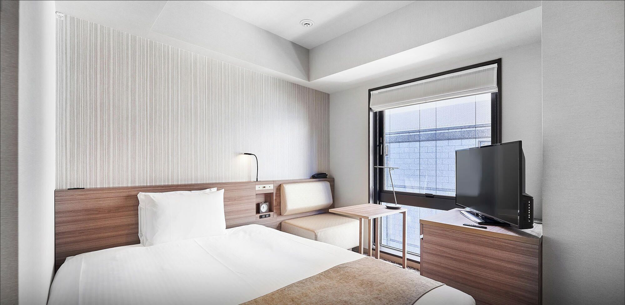 Une chambre d’hôtel moderne, dotée d’un grand lit, d’une télévision, d’un petit bureau et d’une fenêtre donnant sur la ville.