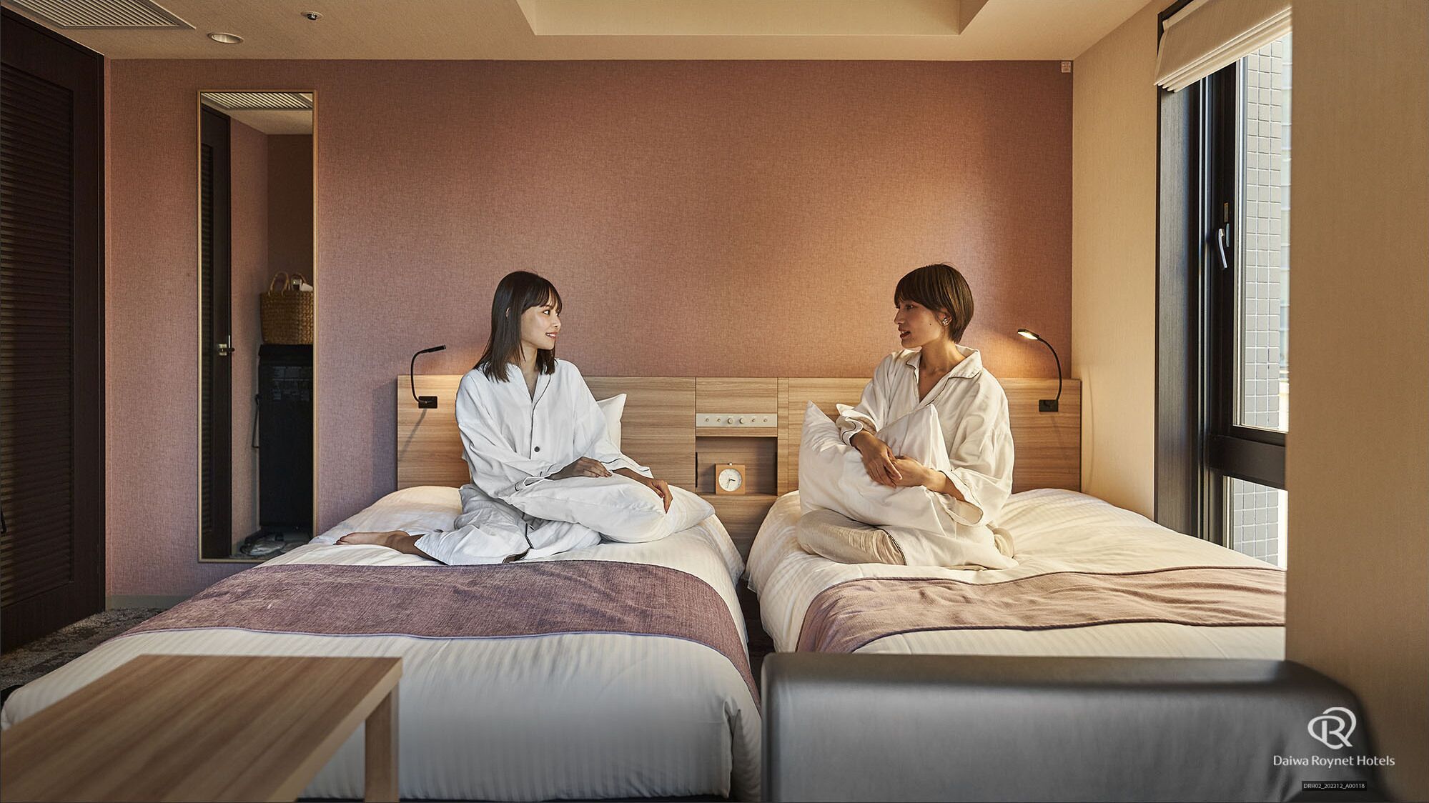 A hotel room with two beds, a wooden headboard, a bedside table, and a small clock.