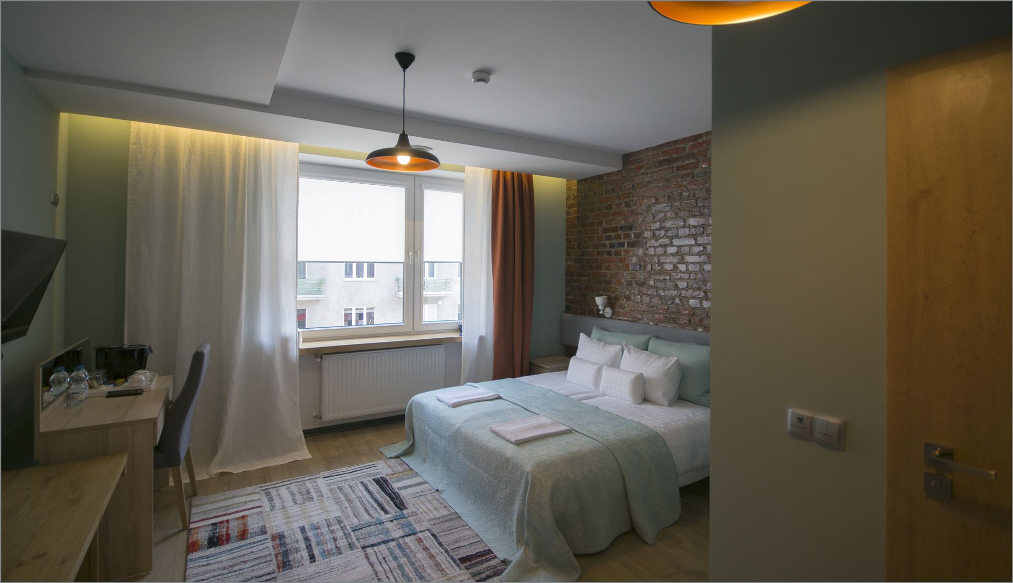 A bedroom with a bed, a desk, a chair, a window with curtains, a radiator, a brick wall, and a hanging light fixture.