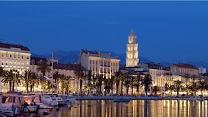 Una veduta della città con lungomare, caratterizzata da una torre imponente, acque calme che riflettono gli edifici e un filare di palme lungo il passeggiata.