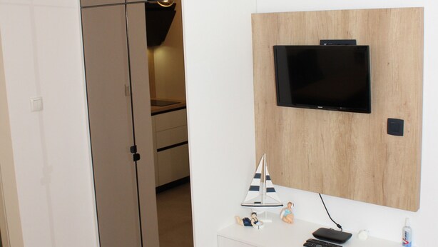 Una stanza moderna con una TV montata su un pannello di legno, un mobiletto bianco con un telecomando e un giocattolo, e una porta vetrata che dà su un'altra stanza.