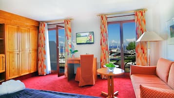 Habitación de hotel con cama, sofá, escritorio con silla, televisión y vistas a un parque y barcos desde las ventanas.