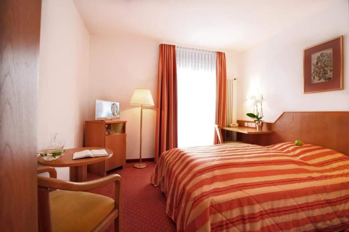 A hotel room with a bed, a desk, a chair, a lamp, and a window with curtains.