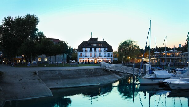 A waterfront hotel with a large building, a dock with boats, and a small bridge. - Schiff am See (Konstanz)