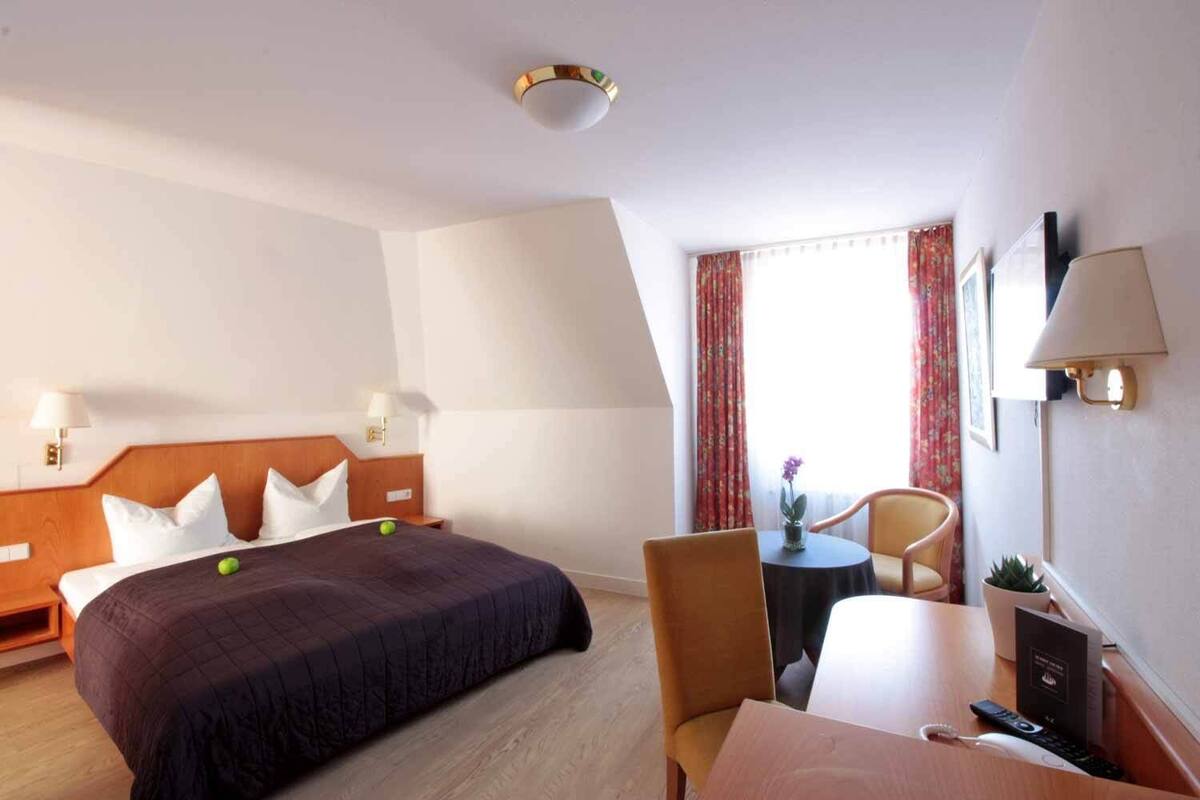 A hotel room with a bed, a desk, a chair, a lamp, and a window with curtains.