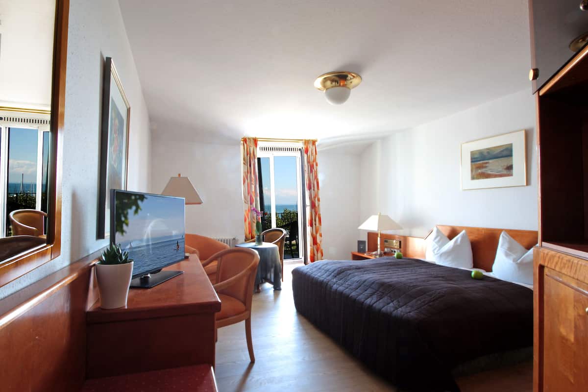 A hotel room with a bed, desk, chair, and a view of the sea through the window.