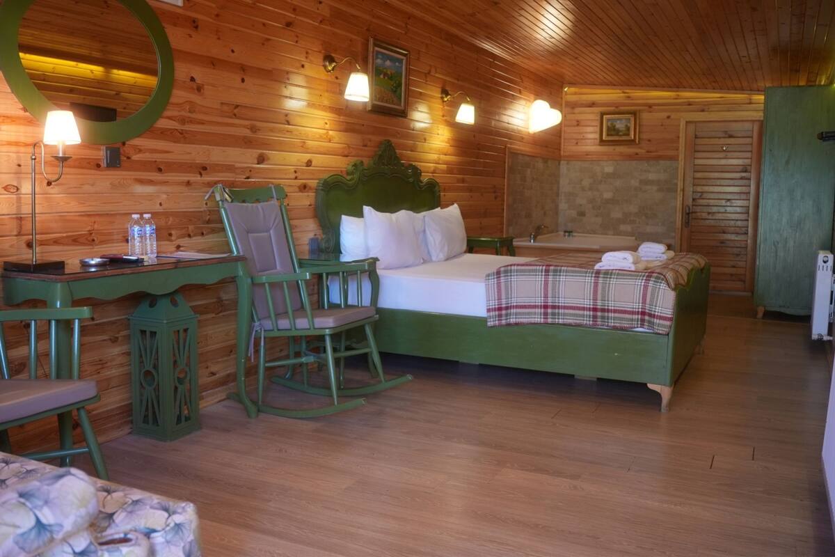 Une pièce de style cabane en bois comprenant un lit, deux chaises, un bureau, un miroir et une salle de bain avec un lavabo et un porte-serviettes.