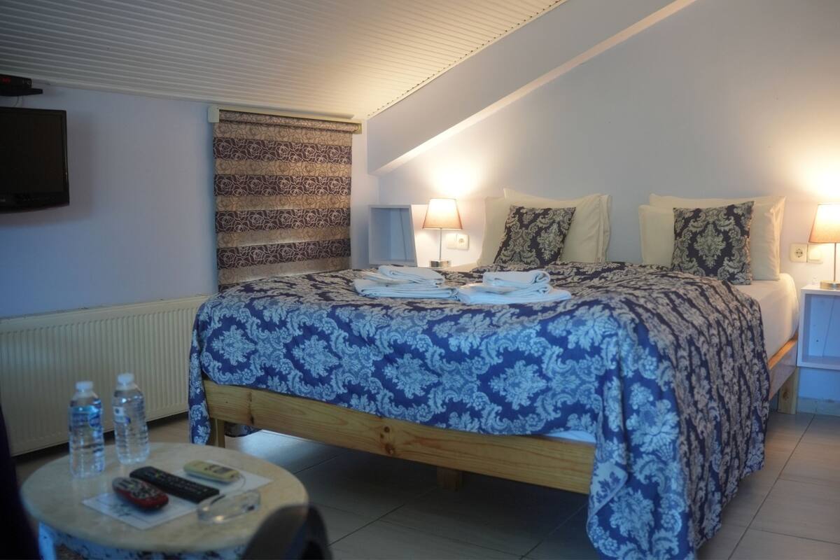 Une chambre avec un lit, deux lampes de chevet, un radiateur, une télévision et une petite table sur laquelle se trouvent des bouteilles d’eau et des télécommandes.