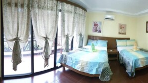 Une chambre d’hôtel avec deux lits, une grande fenêtre avec des rideaux, un ventilateur de plafond et des œuvres d’art accrochées aux murs.