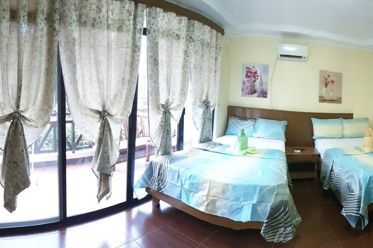 Habitación de hotel con dos camas, un ventanal con cortinas, ventilador de techo y obras de arte en las paredes.