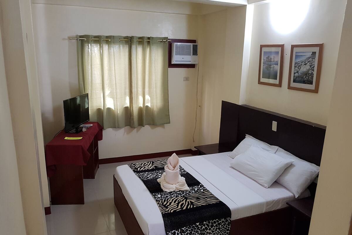 Una habitación de hotel con cama, escritorio, televisión y ventana con cortinas.