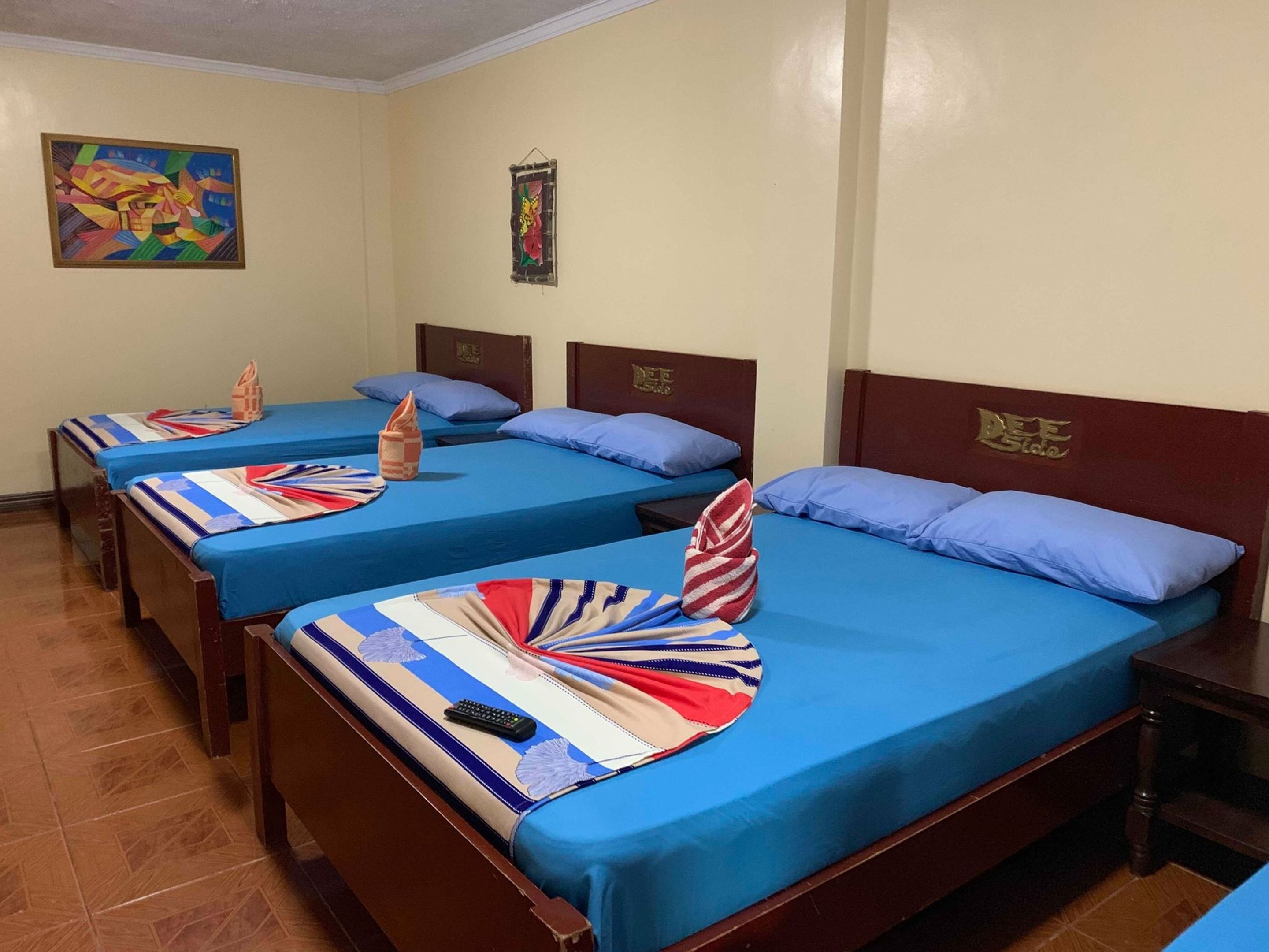 Habitación de hotel con tres camas, cada una con ropa de cama azul y almohadas decorativas. Hay un cuadro en la pared y una mesita con un control remoto en el suelo.