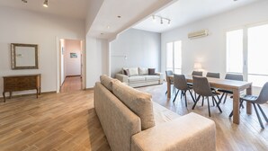 A spacious living area with a sofa, dining table, and chairs. There is a wooden cabinet and a mirror on the wall. - Victoria Majesty Apartments (Athens)