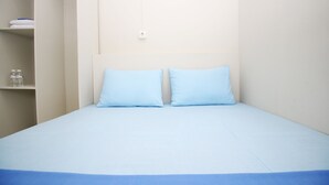 Een kleine eenpersoonskamer met een lichtblauwe spreide en kussens, een witte muur en een plank met opgevouwen handdoeken en flessen.