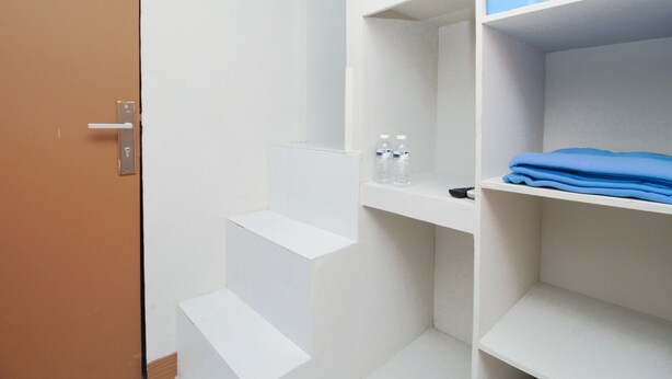 A hotel room with white walls, a brown door, and built-in shelves with a blue container and folded clothes.