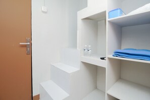 A hotel room with white walls, a brown door, and built-in shelves with a blue container and folded clothes.