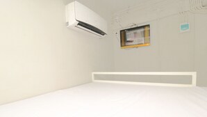 A minimalist bedroom with a white bed, an air conditioning unit, and a small window with a picture frame.