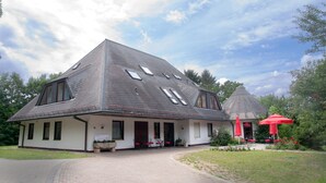 Ein großes, zweistöckiges Gebäude mit geneigtem Dach, zahlreichen Fenstern und einer überdachten Außenbestuhlung mit roten Sonnenschirmen.