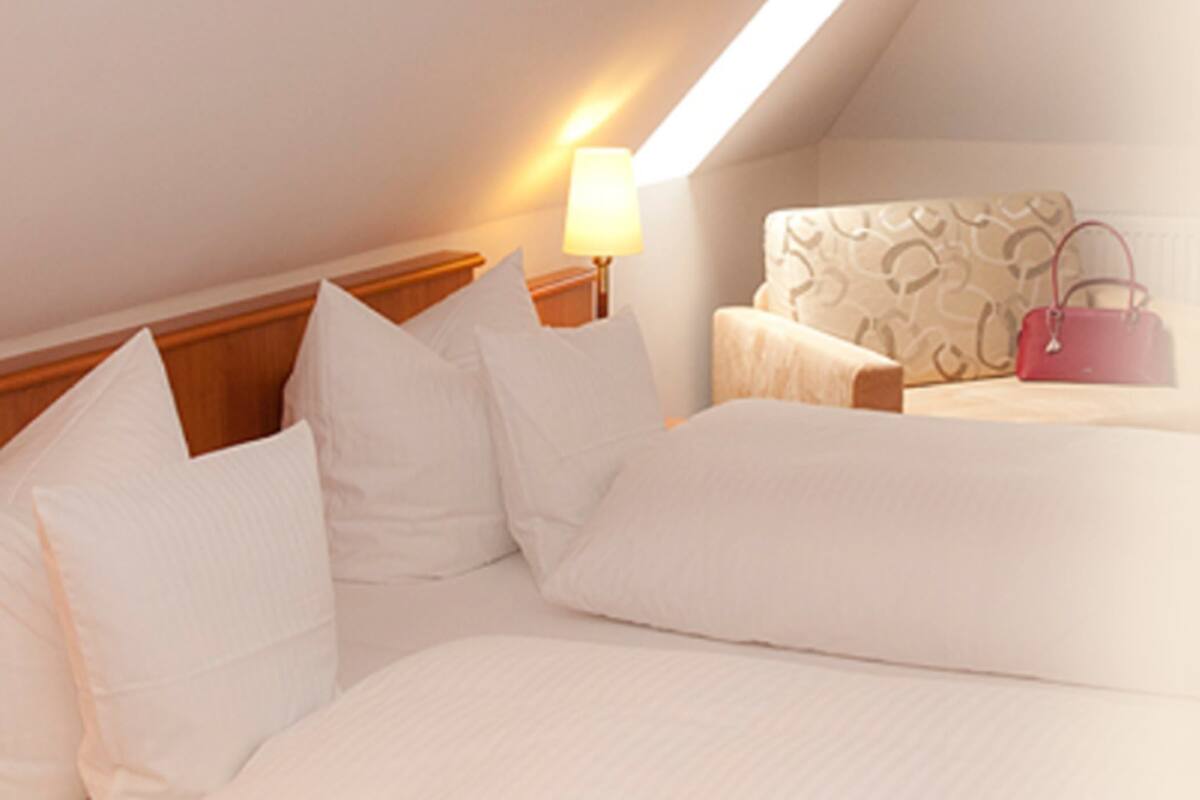 Una habitación de hotel con una cama, almohadas, una mesita de noche, una lámpara, una silla y un bolso.