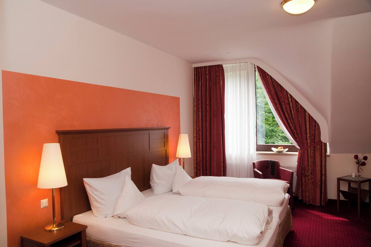 Habitación de hotel con dos camas, cabecera de madera, mesita de noche con lámpara, una silla, una pequeña mesa con un jarrón y una ventana con cortinas.