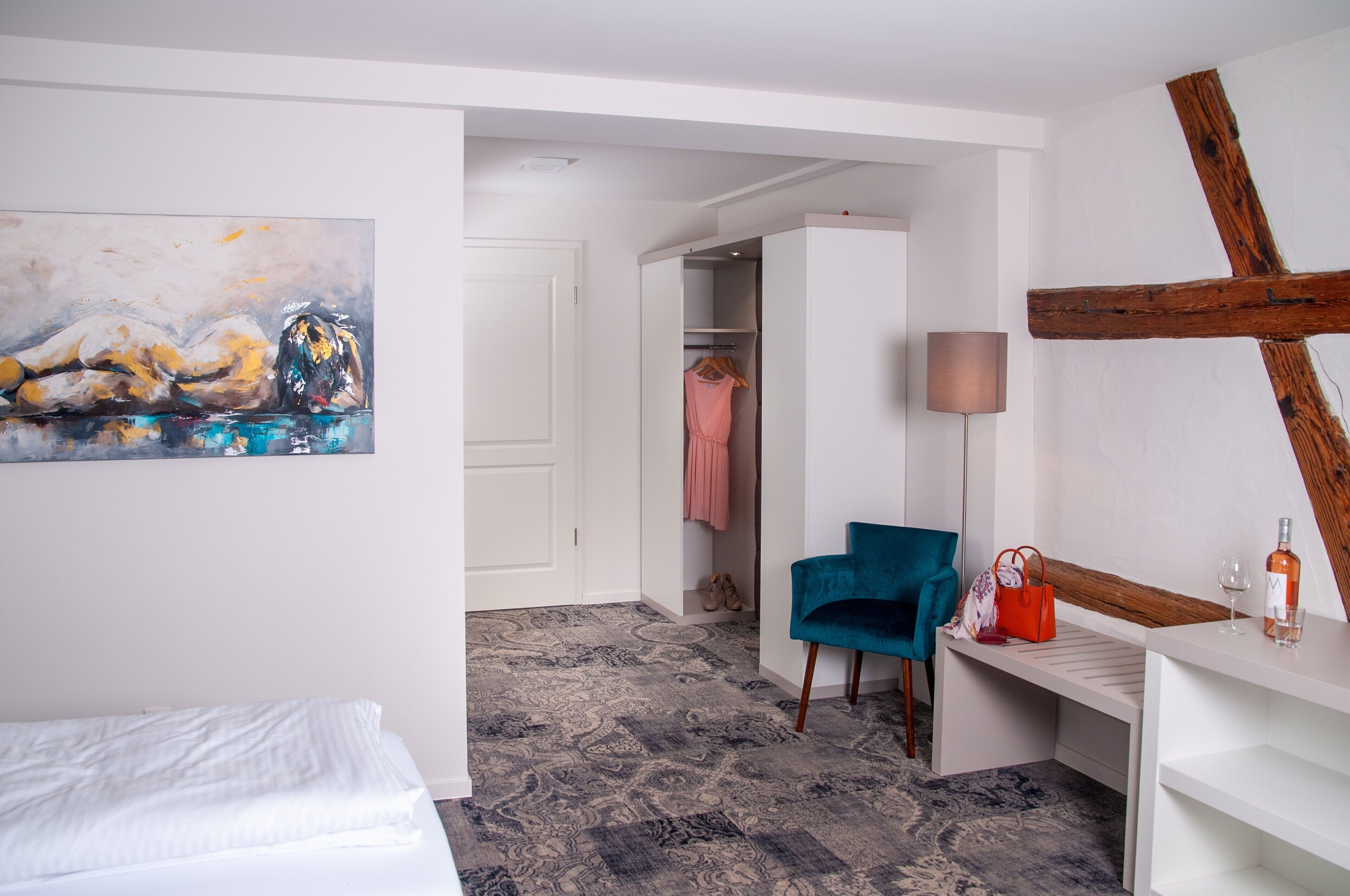 A hotel room with a bed, a desk with a lamp, a chair, a wardrobe, and a painting on the wall.