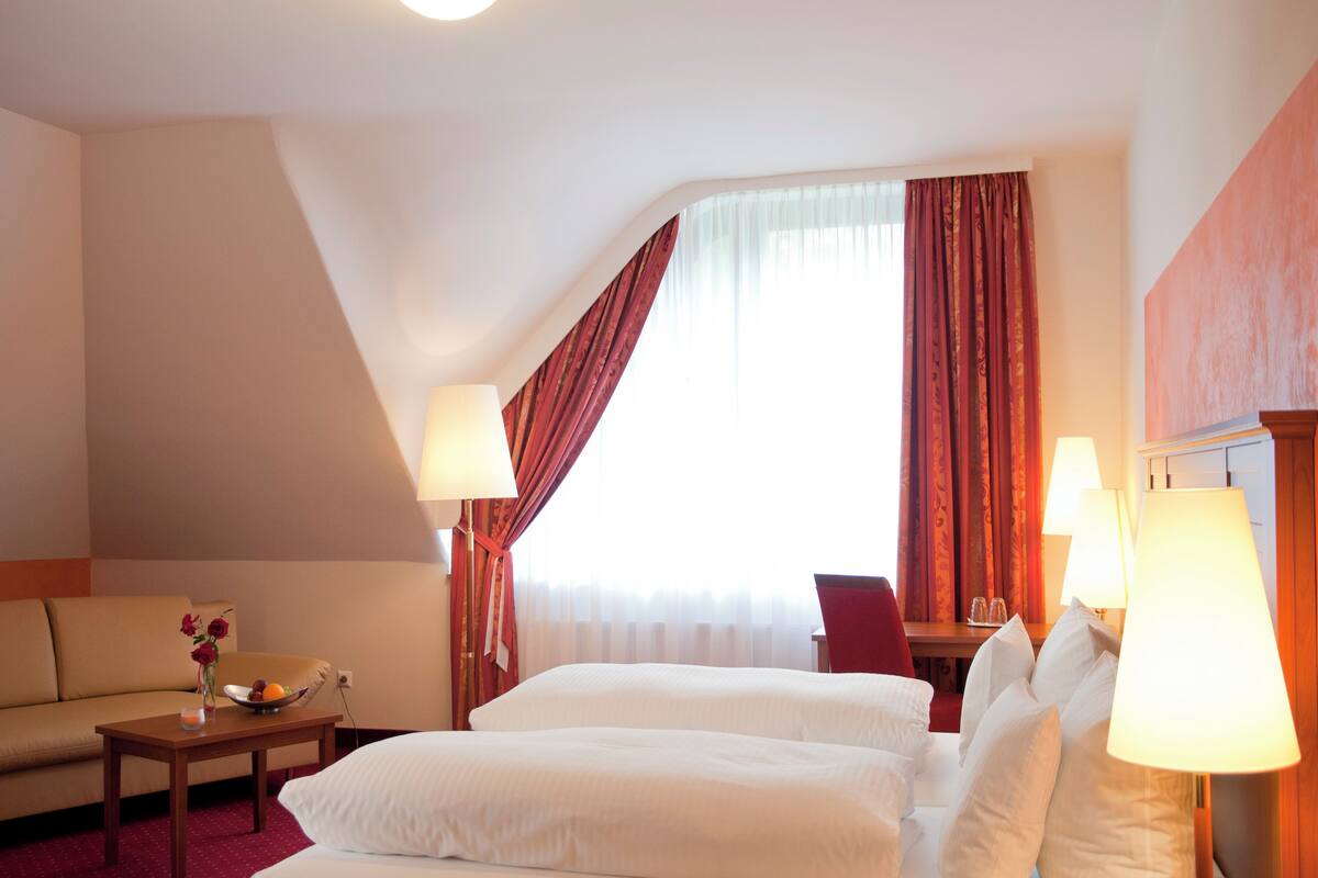 Habitación de hotel con dos camas, un sofá, una mesita con un florero y una ventana con cortinas.