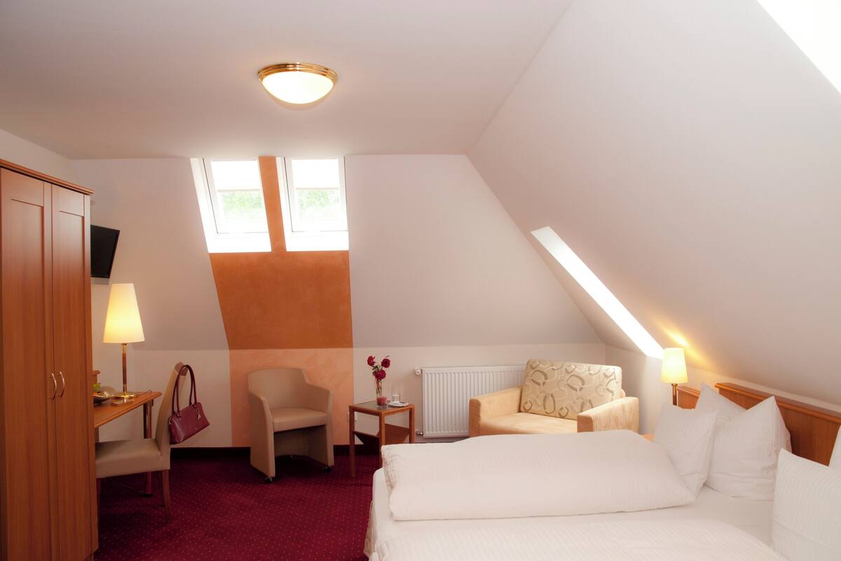 Habitación de hotel con una cama, dos sillas, un escritorio con lámpara, una mesita con un jarrón de flores y una ventana que deja entrar luz natural.