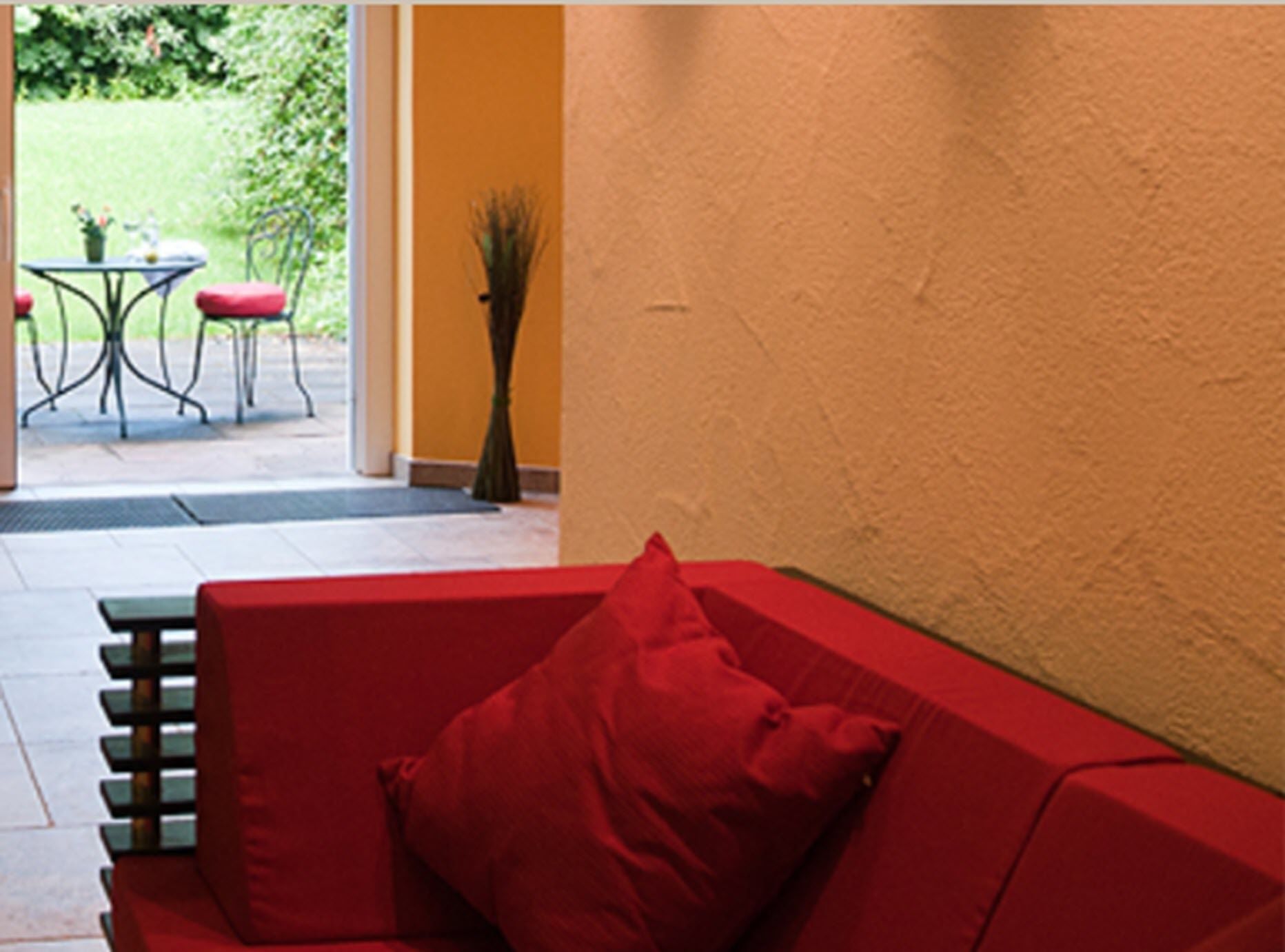 Een rode bank met een rood kussen in een kamer met een gele wand en uitzicht op een terras met een tafel en stoelen.