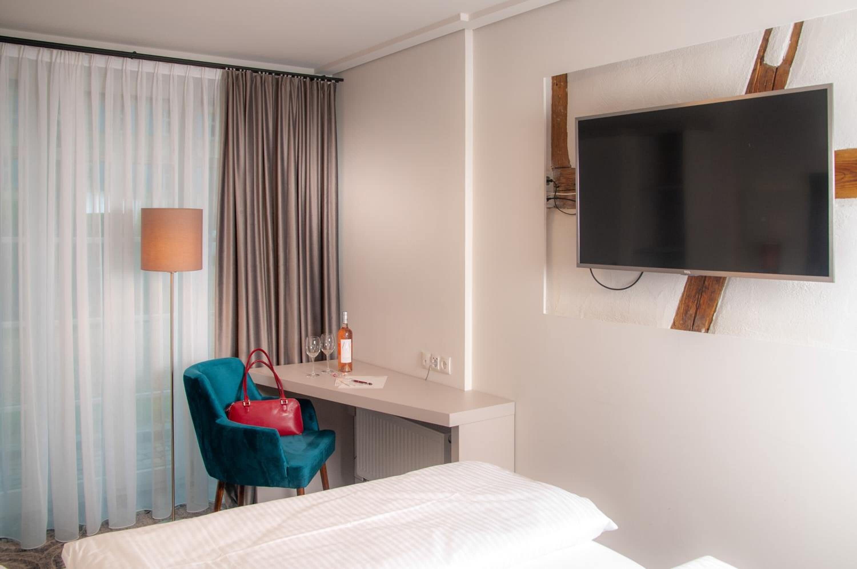Una habitación de hotel con cama, un escritorio con silla, un televisor en la pared y una ventana con cortinas.