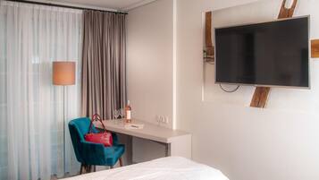 Una habitación de hotel con cama, un escritorio con silla, un televisor en la pared y una ventana con cortinas.