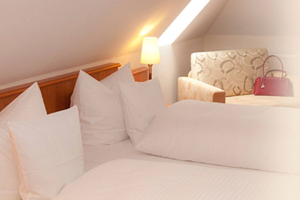 Habitación de hotel con techo inclinado, cama con ropa de cama blanca, mesita de noche con lámpara y un sillón con estampado.