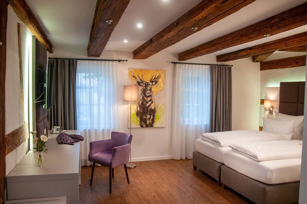 Habitación de hotel con cama, una silla, un cuadro, una lámpara y vigas de madera en el techo.