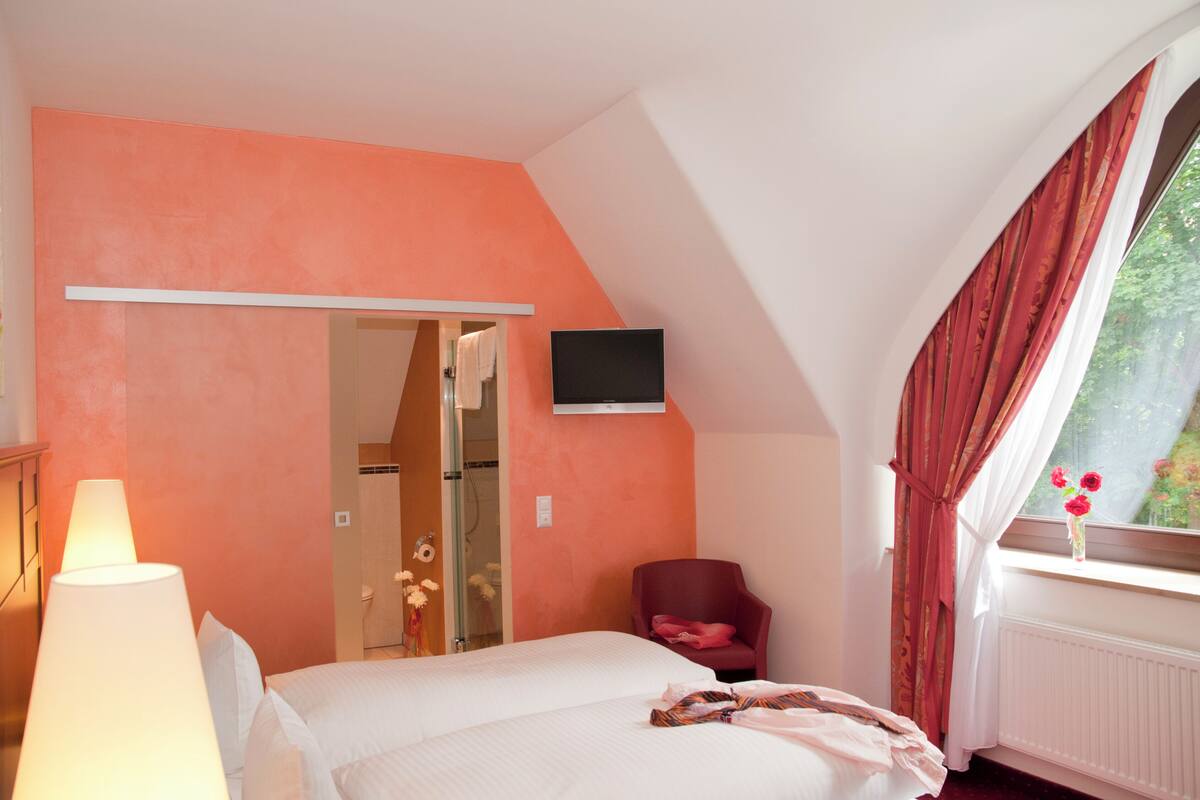 Un dormitorio con cama, televisión montada en la pared, una silla, ventana con cortinas y lámpara.