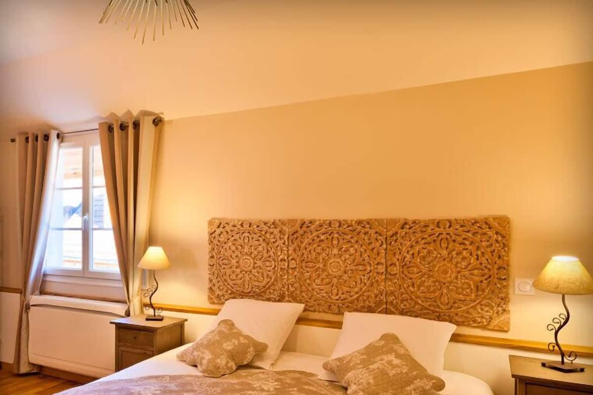 Een keurig opgemaakte slaapkamer met een groot bed, twee nachtkastjes met lampen, een decoratief hoofdeinde en een raam met gordijnen.