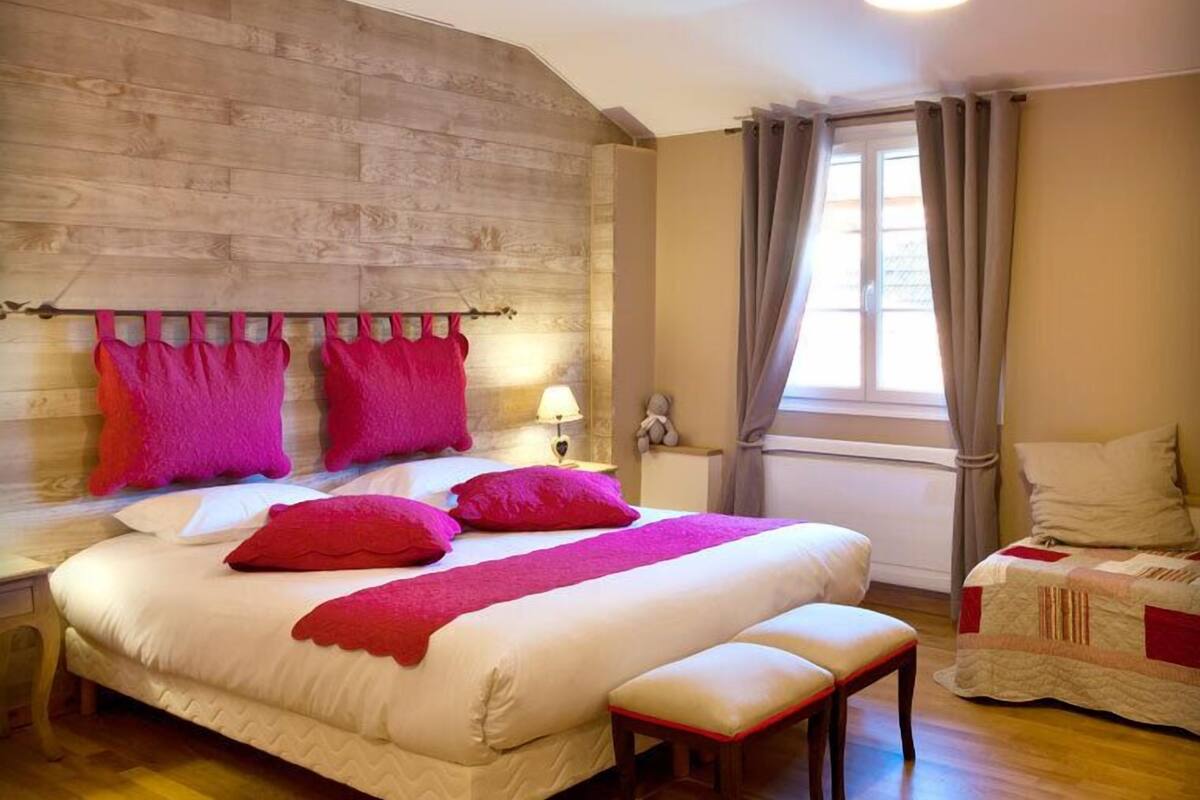 Een slaapkamer met een houten hoofdbord, een bed met roze beddengoed, een bankje, een nachtkastje, een lamp en een raam met gordijnen.
