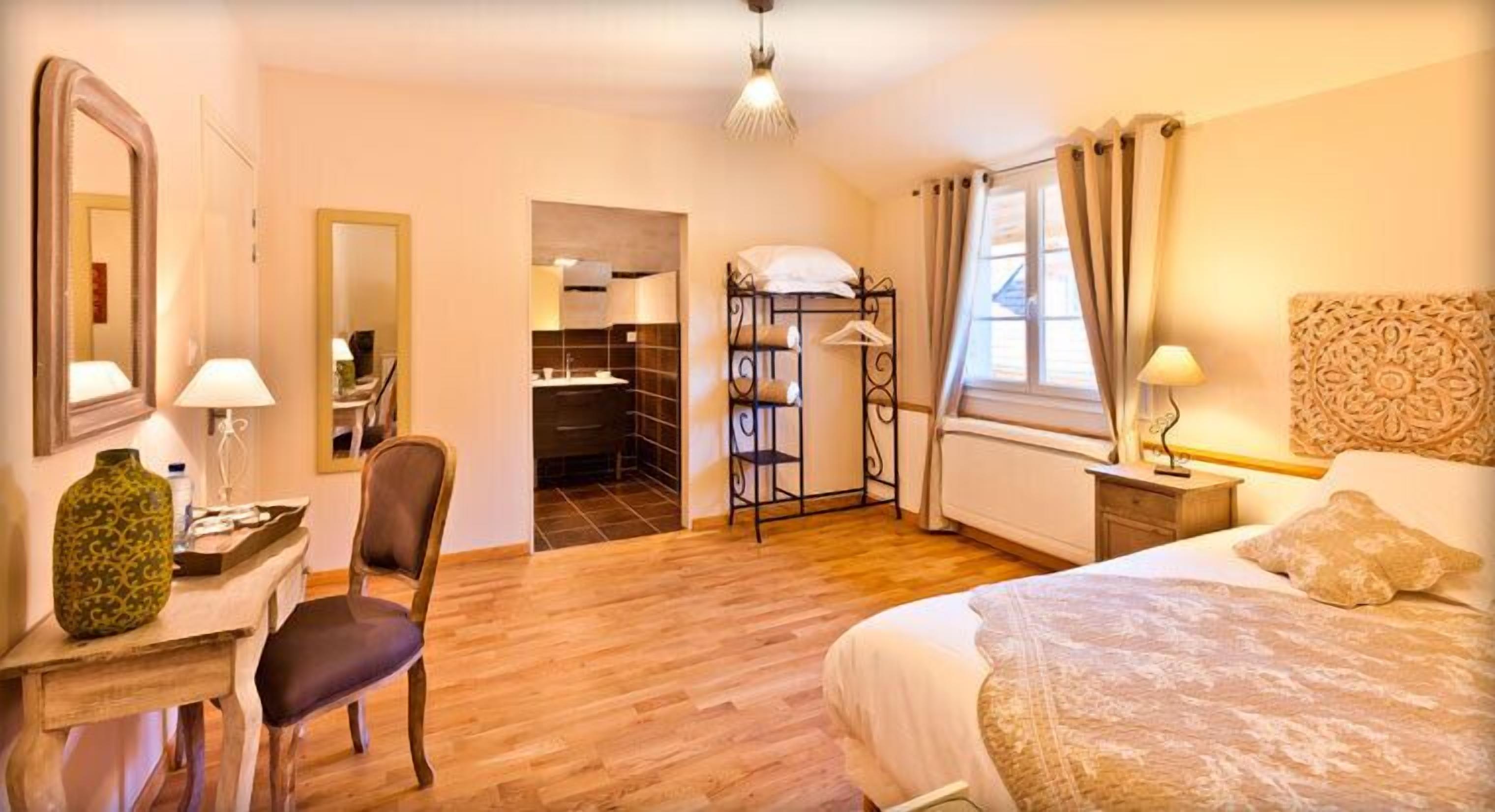 A hotel room with a bed, a desk with a chair, a mirror, a wardrobe, and a bathroom visible through an open door.