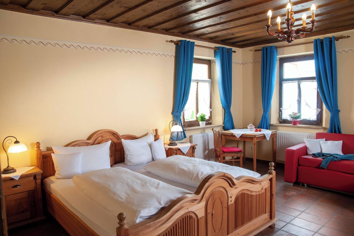 A bedroom with a wooden bed, a red sofa, a small table with a lamp, and a window with blue curtains.