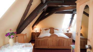 A bedroom with a wooden bed, a bedside table with a lamp, a small table with a vase of flowers, and a door leading to another room.