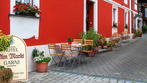 A red building with a sign for Hotel Im Markt, featuring outdoor seating with wooden chairs and tables, potted plants, and a cobblestone street.